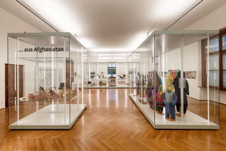 A spacious exhibition hall featuring glass display cases showcasing various artifacts and clothing items from Afghanistan. The wooden floor complements the modern design, and there is a prominent sign reading