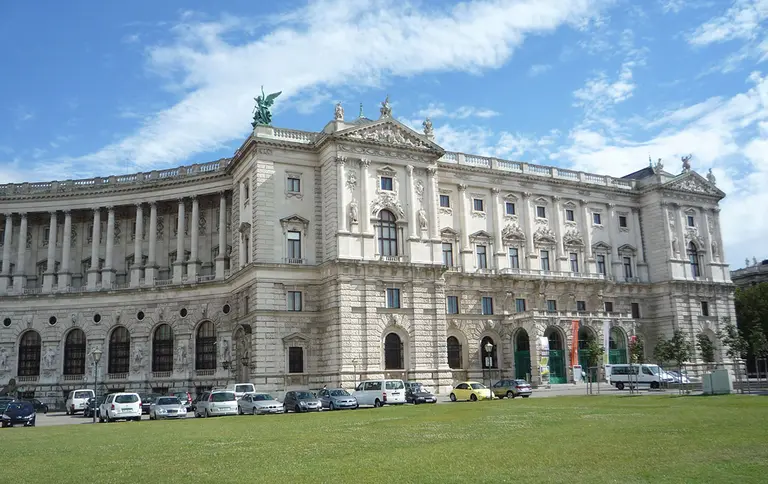 A grand, historic building with intricate architecture, featuring large columns and decorative details. The façade is adorned with statues, and green grass is in the foreground, with several parked cars along the side. The sky is partly cloudy, creating a vibrant backdrop.