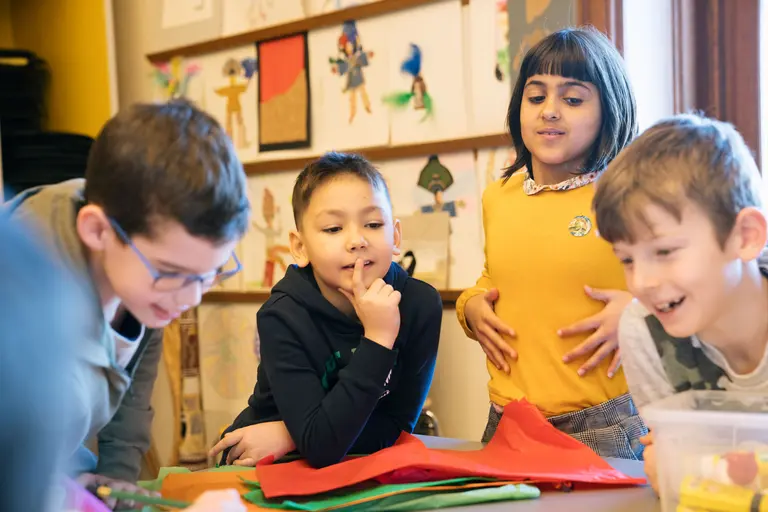 Fünf Kinder stehen um einen Tisch, auf dem farbiges Papier liegt. Sie lächeln und scheinen an einem kreativen Projekt zu arbeiten. Ein Junge hat den Finger an seine Lippen gelegt, während ein anderes Kind aufgeregt gestikuliert. Die Atmosphäre ist fröhlich und beschäftigts.
