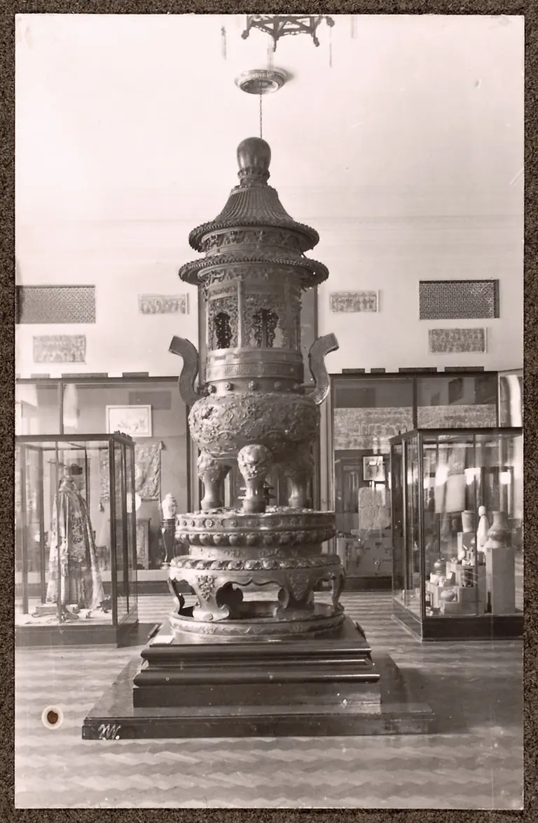 A large, ornate bronze incense burner stands in the center of a museum exhibit. It features intricate carvings, including figures and decorative motifs, and is displayed on a dark pedestal. Display cases showcasing various artifacts surround it, with soft lighting illuminating the piece.