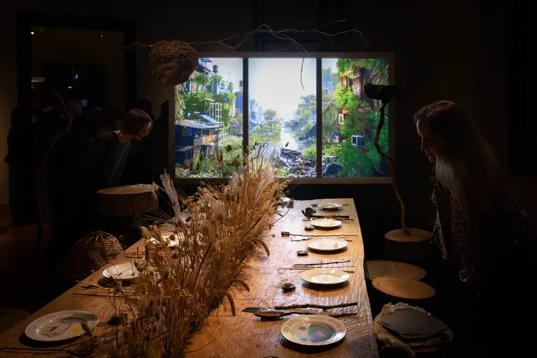 Ein gedeckter Tisch mit Tellern und trockenem Gras, vor einem großen Bildschirm, der eine grüne Landschaft zeigt. Menschen stehen am Tisch und betrachten die Szene. Der Raum ist schwach beleuchtet und die Atmosphäre ist einladend.