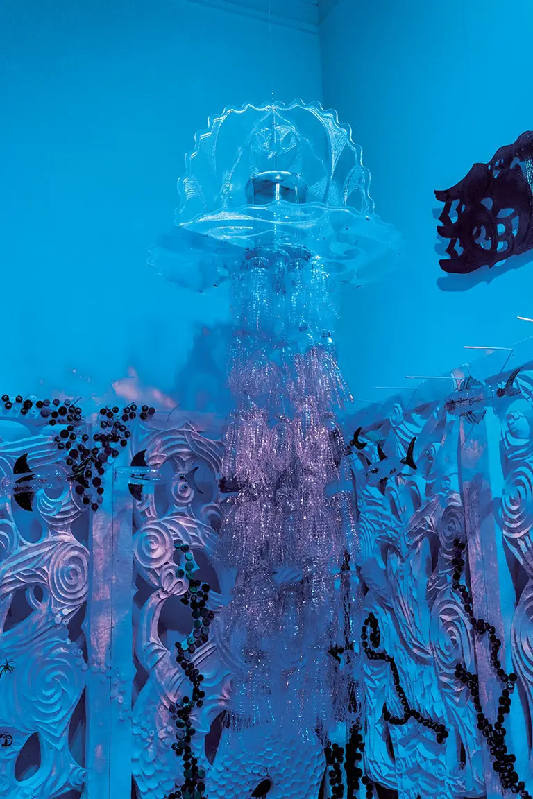 A large, transparent jellyfish sculpture hangs from the ceiling, illuminated in blue light. The sculpture has a domed top and flowing, beaded tentacles that create a shimmering effect, set against a backdrop of textured, organic patterns in a similar color scheme.