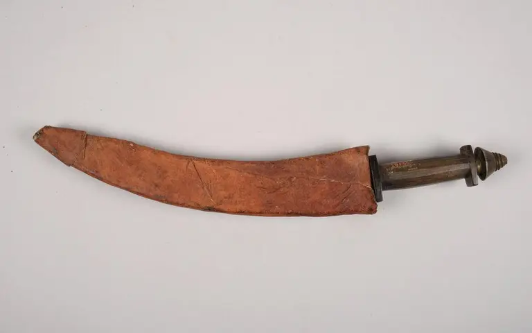 A curved, traditional dagger is displayed. The blade is partially covered by a distinct leather sheath, which has a smooth surface and a tapered end. The handle features ornate details, showing cultural craftsmanship. The overall appearance reflects historical significance and artistry in weapon design.