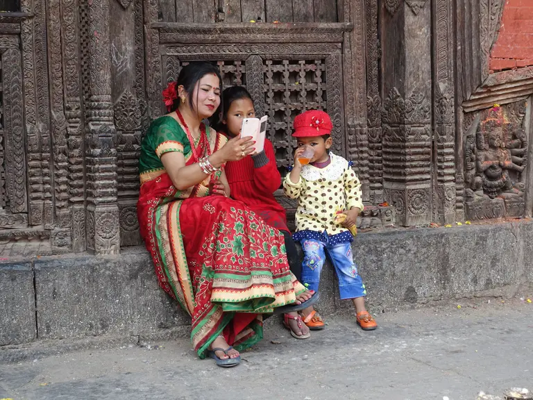 A woman in traditional attire sits on a stone step, sharing a moment with two children. One child, wearing a red sweater, looks at a smartphone, while the younger child, dressed in a colorful outfit and cap, observes. They are in front of an intricately carved wall.