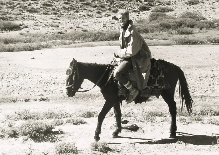 A person riding a horse through a rugged landscape, wearing a long coat and boots. The setting is a dry, open area with sparse vegetation in the background, captured in black and white.