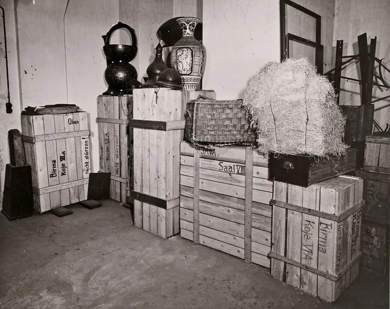 A collection of wooden crates and boxes stacked against a wall in a dimly lit room. Some crates are labeled with various names, while decorative pottery and a woven basket are placed on top of the crates, adding to the scene's vintage atmosphere.