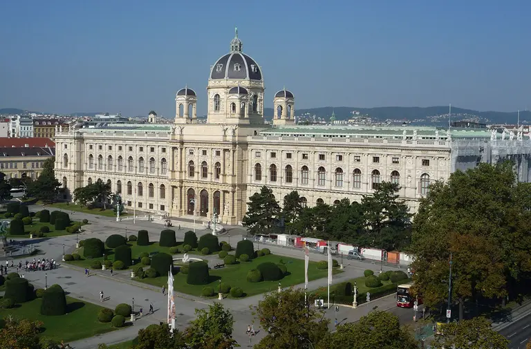 A large historic building with a prominent dome and decorative architecture, surrounded by manicured gardens and trees. The scene is set against a clear blue sky, showcasing the grandeur of the structure and its landscaped surroundings.