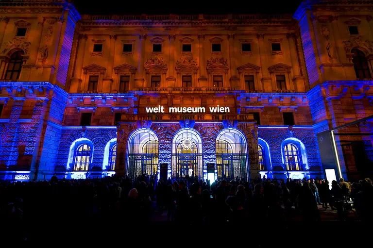 The illuminated facade of the Welt Museum Wien at night, featuring a blend of blue and orange lighting. A large crowd gathers in front, with the museum's name displayed prominently above the entrance.