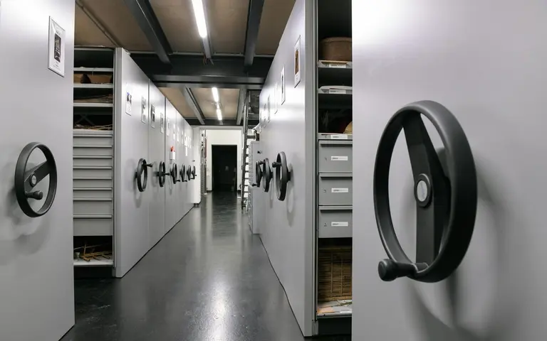The image shows a long corridor lined with storage cabinets equipped with large, black hand-crank handles. The cabinets are gray, and a doorway is visible at the end of the hallway, leading to a darker space. The floor is smooth and dark.